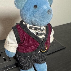 May include: A blue teddy bear figurine wearing a burgundy vest, black and white striped scarf, and gray patterned pants. The bear has a gold-colored ring on the vest and is standing on a black surface. The bear's ears and feet are also blue.