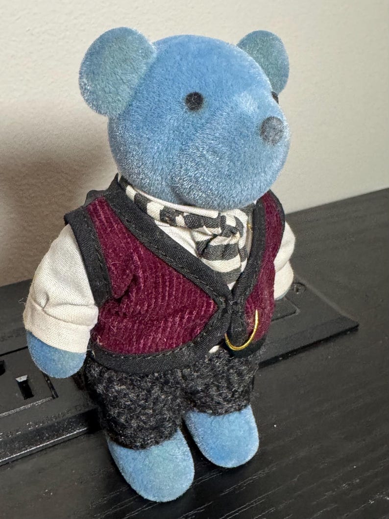 May include: A blue teddy bear figurine wearing a burgundy vest, black and white striped scarf, and gray patterned pants. The bear has a gold-colored ring on the vest and is standing on a black surface. The bear's ears and feet are also blue.