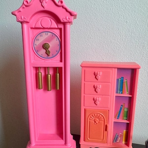 Könnte beinhalten: Rosa Spielzeuguhr und Schrank-Set. Die Uhr hat ein rundes Zifferblatt mit goldfarbenen Zeigern und Pendel. Der Schrank hat Schubladen und eine Tür mit Totenkopf-Design. Der Schrank hat ein Bücherregal mit Miniaturbüchern.
