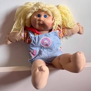 Könnte beinhalten: Eine Vintage-Cabbage-Patch-Kids-Puppe mit blondem Garnhaar in Zöpfen. Die Puppe hat blaue Augen, rosige Wangen und trägt ein Hemd mit Blumenmuster unter einer blauen Latzhose mit einem rosa und blauen Aufnäher.