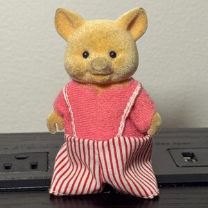 May include: A vintage toy pig figurine with a yellow head and a pink outfit. The pig is wearing a pink top with white straps and red and white striped pants. The toy has black eyes and a smiling expression.