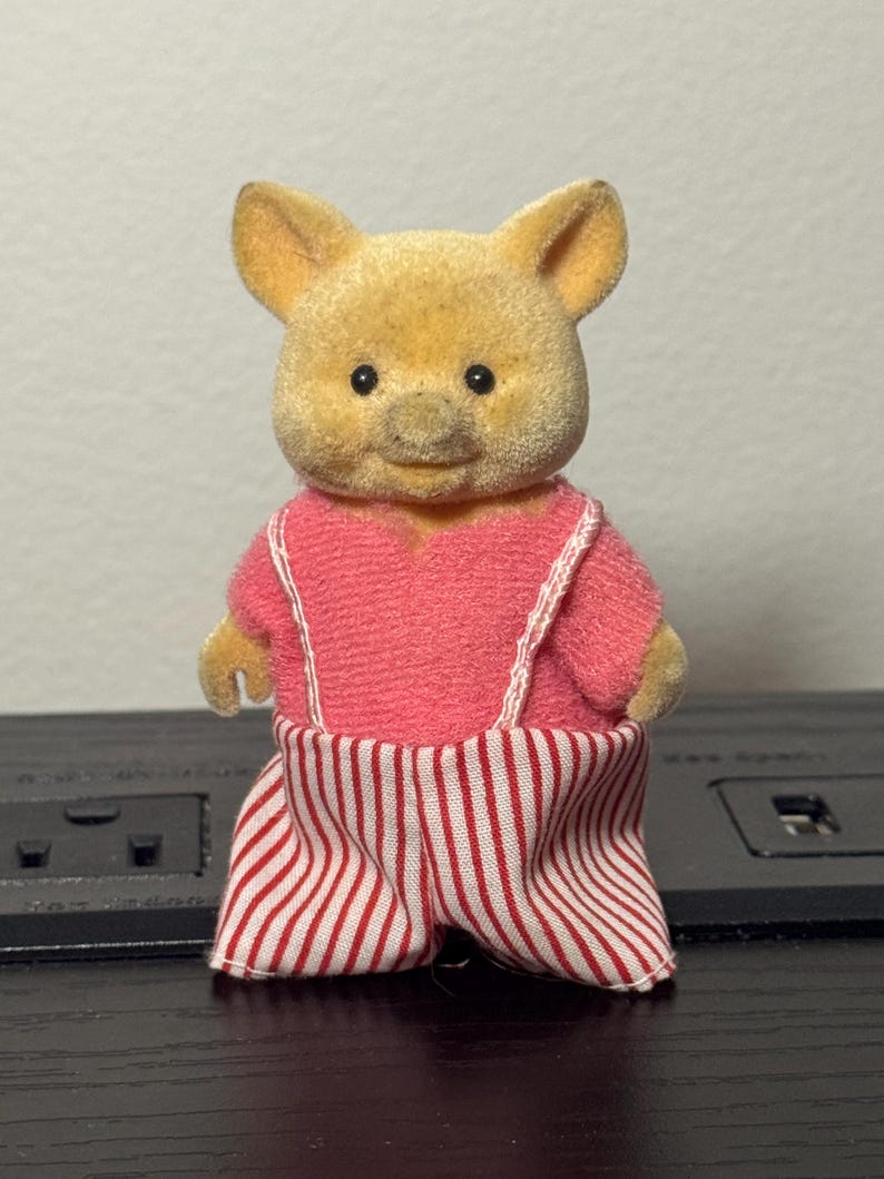 May include: A vintage toy pig figurine with a yellow head and a pink outfit. The pig is wearing a pink top with white straps and red and white striped pants. The toy has black eyes and a smiling expression.