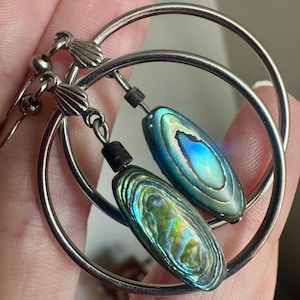 May include: A pair of silver-toned hoop earrings featuring dangling, elongated oval-shaped abalone shell beads. Each bead displays iridescent blue and green hues with a natural, polished texture. The earrings are attached to silver-toned metal hooks.
