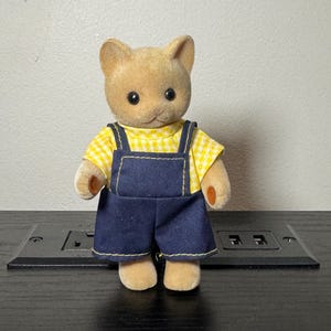 May include: A tan cat figurine wearing a yellow gingham shirt and blue denim overalls. The figurine has black eyes and small, tan paws. The overalls have yellow stitching.