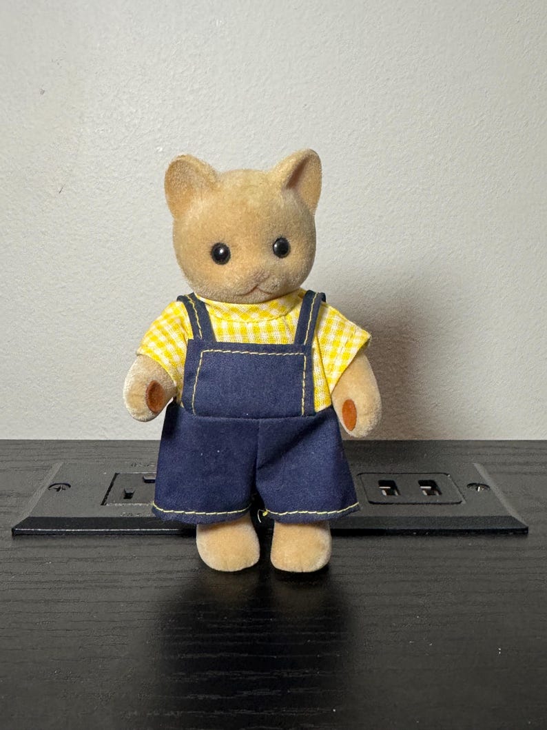 May include: A tan cat figurine wearing a yellow gingham shirt and blue denim overalls. The figurine has black eyes and small, tan paws. The overalls have yellow stitching.