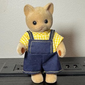 May include: A tan cat figurine wearing a yellow and white checkered shirt and dark blue overalls. The figurine has black eyes and small, tan paws. The overalls have yellow stitching.