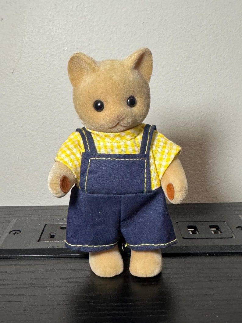 May include: A tan cat figurine wearing a yellow and white checkered shirt and dark blue overalls. The figurine has black eyes and small, tan paws. The overalls have yellow stitching.