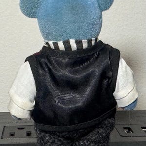 May include: A blue teddy bear wearing a black vest over a white shirt with a black and white striped collar. The bear also has gray patterned shorts. The bear's ears are round and blue.