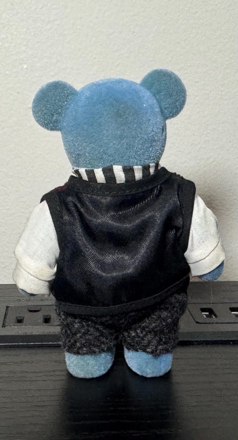 May include: A blue teddy bear wearing a black vest over a white shirt with a black and white striped collar. The bear also has gray patterned shorts. The bear's ears are round and blue.