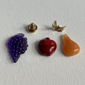 Puede incluir: Un surtido de joyas y abalorios, que incluye un racimo de uvas moradas, una manzana roja y una pera naranja. También se incluyen un perno de color dorado y un abalorio de color dorado.