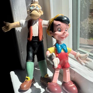 May include: Two moulded plastic figurines depicting Geppetto and Pinocchio. Geppetto wears a white shirt, red waistcoat, black trousers, and green leggings. Pinocchio wears a yellow shirt, blue bow tie, red shorts, and a yellow hat with a black band.