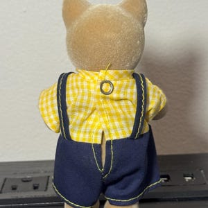 May include: A small, tan-colored animal figurine wearing a yellow and white checkered shirt and dark blue overalls with suspenders. The figurine has a button closure on the back of the shirt.