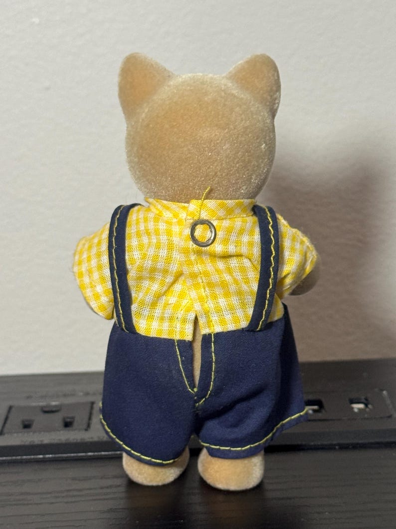 May include: A small, tan-colored animal figurine wearing a yellow and white checkered shirt and dark blue overalls with suspenders. The figurine has a button closure on the back of the shirt.