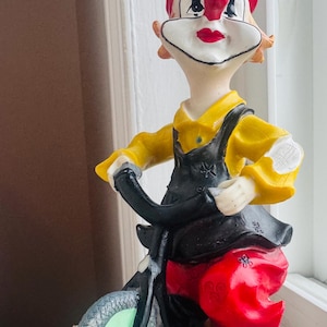 May include: A ceramic clown figurine on a bicycle. The clown has a red hat, yellow shirt, and black overalls. The bicycle has green tyres and a red seat. The figurine is on a white base with yellow flower details.