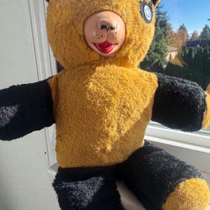Può includere: Un costume vintage da orsetto con corpo giallo e dettagli neri. L'orso ha un viso di plastica con bocca rossa e un solo occhio. Il costume è realizzato in materiale sfocato.