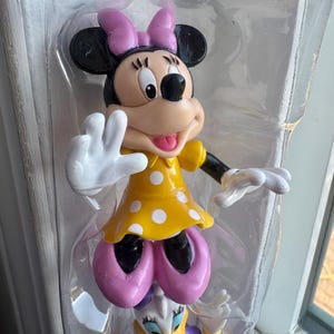 May include: A Minnie Mouse figurine encased in clear plastic packaging, featuring a yellow dress with white polka dots, a pink bow, and pink shoes. The character has black ears, a tan face, and white gloves, with one hand raised in a wave.