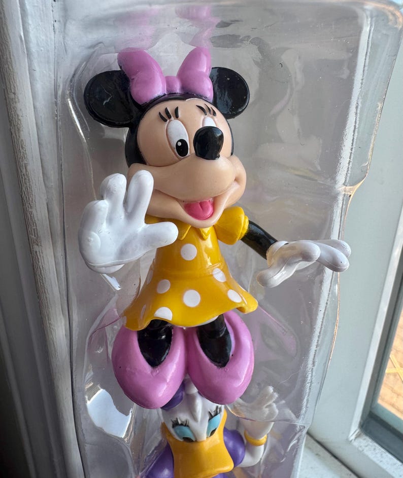 May include: A Minnie Mouse figurine encased in clear plastic packaging, featuring a yellow dress with white polka dots, a pink bow, and pink shoes. The character has black ears, a tan face, and white gloves, with one hand raised in a wave.