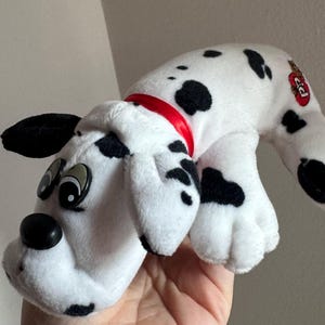 May include: A plush Dalmatian dog toy with a red collar. The toy is white with black spots and has large, cartoonish eyes. The dog is lying down, with its head and front paws visible.