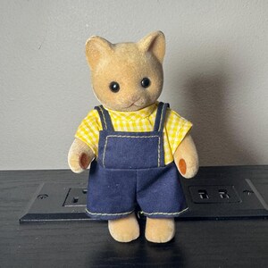 May include: A tan cat figurine wearing a yellow gingham shirt and dark blue denim overalls with yellow stitching. The figurine has black eyes and small, rounded ears. The toy is standing on a black surface.