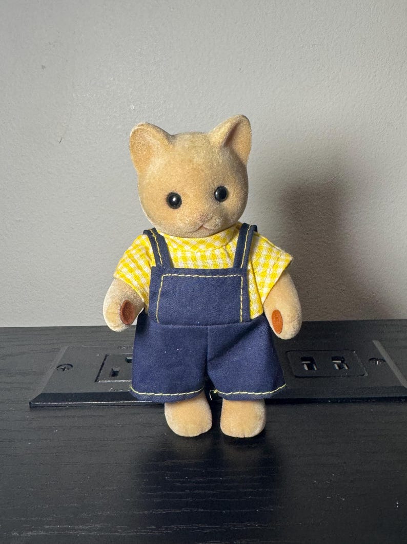 May include: A tan cat figurine wearing a yellow gingham shirt and dark blue denim overalls with yellow stitching. The figurine has black eyes and small, rounded ears. The toy is standing on a black surface.
