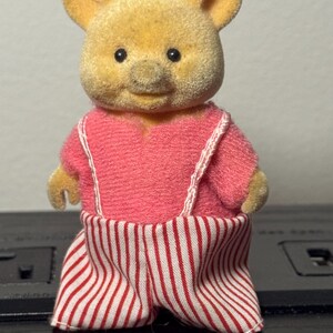 May include: A vintage toy pig figurine with a yellow head and pink outfit. The pig is wearing a pink shirt with white suspenders and red and white striped pants. The toy has black eyes and a smiling expression.