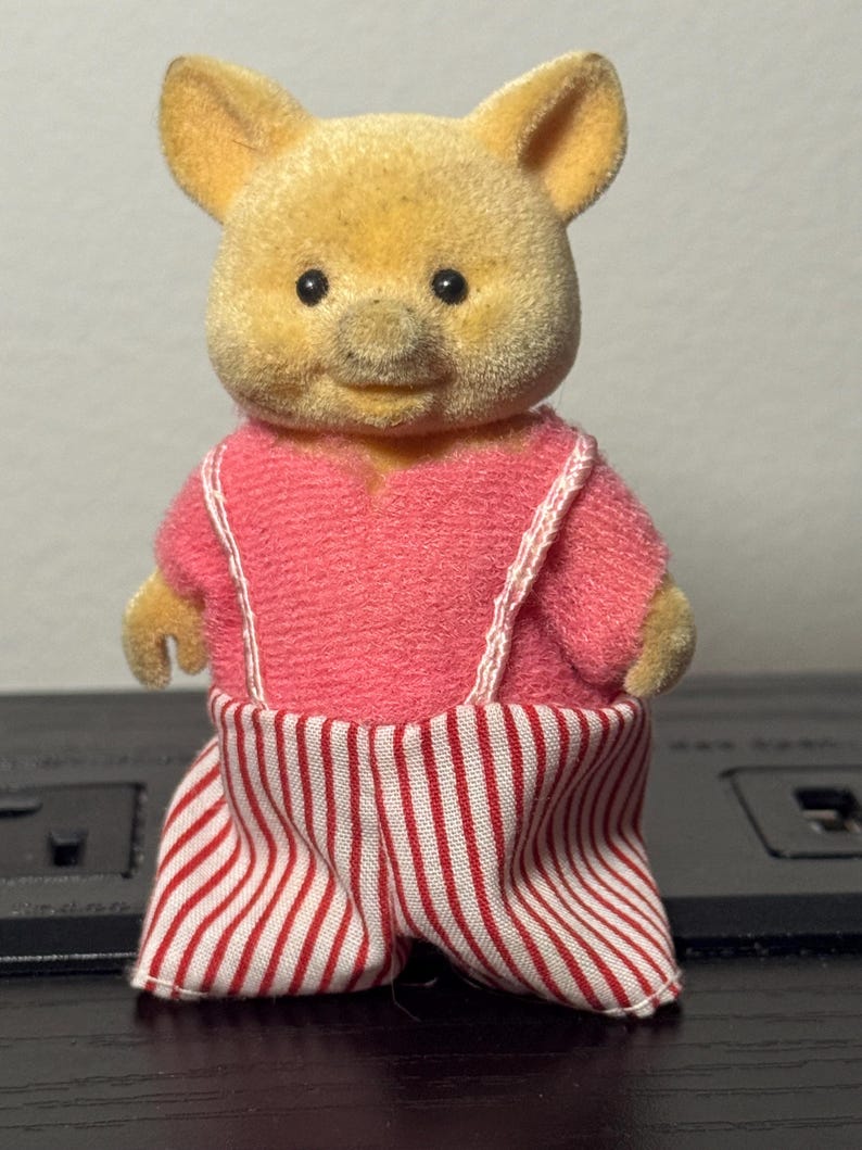 May include: A vintage toy pig figurine with a yellow head and pink outfit. The pig is wearing a pink shirt with white suspenders and red and white striped pants. The toy has black eyes and a smiling expression.