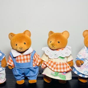May include: Four toy bear figures with brown fur, each dressed in different outfits. One wears a blue and white checkered shirt and pants with a pink bow tie. Another has blue overalls. The other two wear dresses.