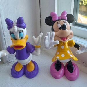 May include: Two plastic character figurines. On the left, a white duck with a yellow beak, purple shirt, purple bow, and purple shoes. On the right, a black mouse with a pink bow, yellow dress with white polka dots, and pink shoes.