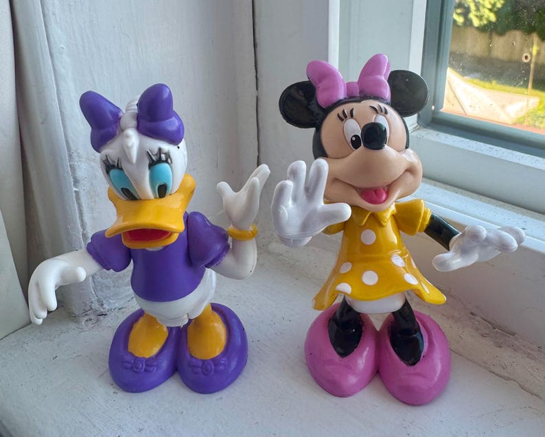 May include: Two plastic character figurines. On the left, a white duck with a yellow beak, purple shirt, purple bow, and purple shoes. On the right, a black mouse with a pink bow, yellow dress with white polka dots, and pink shoes.