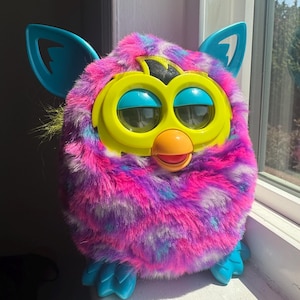 May include: A vibrant Furby toy with a fluffy body in pink, purple, and blue hues. It features large yellow eyes, an orange beak, and blue ears and feet. The toy is positioned on a white windowsill, catching the sunlight.