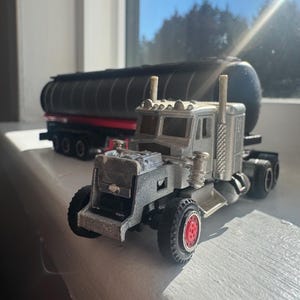 May include: A silver and grey toy articulated lorry with red wheel details. A black tanker truck is in the background. The toy truck is on a white surface near a window.