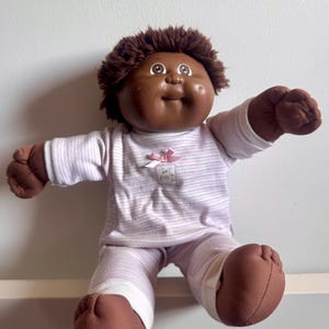 Könnte beinhalten: Eine Vintage-Cabbage-Patch-Kid-Puppe mit brauner Haut, braunen Haaren und braunen Augen. Die Puppe trägt einen hellviolett-weiß gestreiften Pyjama mit einer rosa Schleife. Die Puppe sitzt mit ausgestreckten Armen.