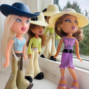 May include: Four Bratz dolls are displayed, each with unique outfits and accessories. One doll wears a blue cowboy hat, a blue top, and olive green pants. Another doll is in a light green top and white pants. The third doll is in a purple dress and a wide-brimmed hat.