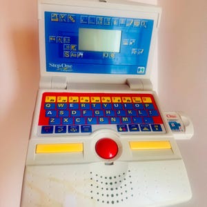 May include: A white and blue Step One Compact computer toy with a red, yellow, and blue keyboard. The toy has a small screen and a red trackball. The text "Step One Computers" is visible.