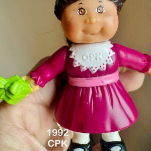 Puede incluir: Muñeca vintage Cabbage Patch Kids con cabello castaño y un vestido rosa. La muñeca tiene zapatos negros y un cuello blanco con "CPK" impreso. El año "1992" y "CPK MCD" están impresos en la muñeca.