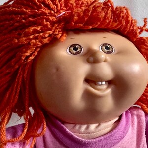 Puede incluir: Una muñeca Cabbage Patch Kid vintage con cabello de hilo naranja y ojos marrones. La muñeca tiene una expresión sonriente y lleva un atuendo rosa y morado. La cara de la muñeca es de tono claro.