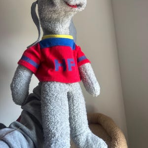May include: A plush, gray meerkat-like stuffed animal wearing a red shirt with blue and yellow trim. The shirt has the letters "HP" in blue. The toy has a long, gray tail and ears.