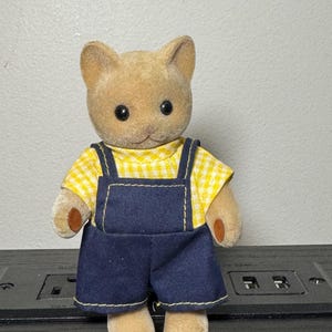 May include: A small, plush cat figurine wearing a yellow gingham shirt and dark blue denim overalls. The cat has a light brown fur coat, black eyes, and small brown paws. The overalls have yellow stitching.