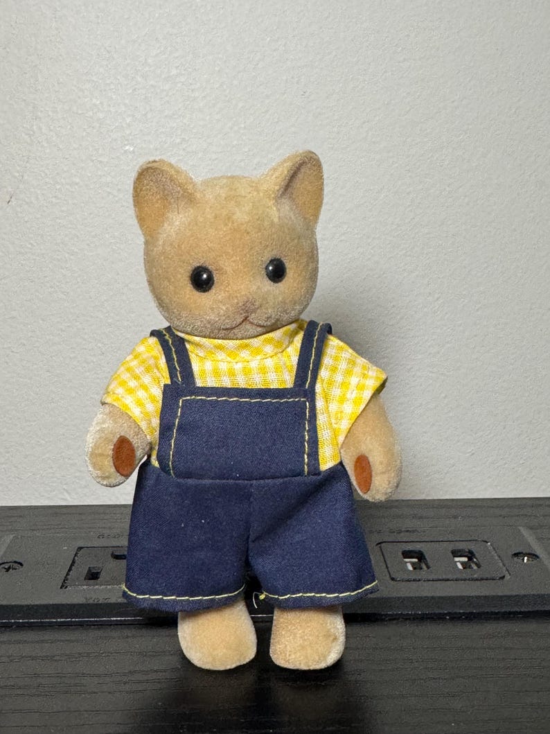May include: A small, plush cat figurine wearing a yellow gingham shirt and dark blue denim overalls. The cat has a light brown fur coat, black eyes, and small brown paws. The overalls have yellow stitching.