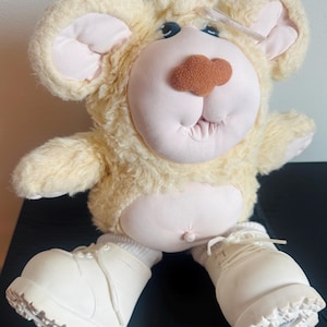 Pode incluir: Um urso de pelúcia com um corpo de pele amarelo claro, orelhas grandes e um rosto rosa. O urso tem botas brancas e um nariz castanho. O brinquedo é projetado com um estilo fantasioso e caricatural.