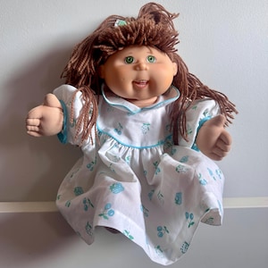 Könnte beinhalten: Eine Vintage Cabbage Patch Kid Puppe mit braunem Garnhaar und grünen Augen. Die Puppe trägt ein weißes Kleid mit blauer Bordüre und einem Muster aus blauen Beeren und Blättern. Die Puppe hat einen lächelnden Ausdruck.