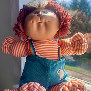 Op de afbeelding: Een vintage Cabbage Patch Kid-pop met rood garen haar en blauwe ogen. De pop draagt een gestreept oranje en wit shirt, een blauwe corduroy overall met regenboog bretels en bijpassende gestreepte voeten.