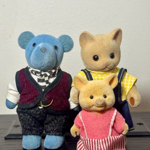 May include: Three miniature animal figurines: a blue bear, a tan cat, and a yellow pig. The bear wears a burgundy vest and checkered scarf. The cat has a yellow top with blue overalls. The pig is in a pink dress with red and white striped pants.