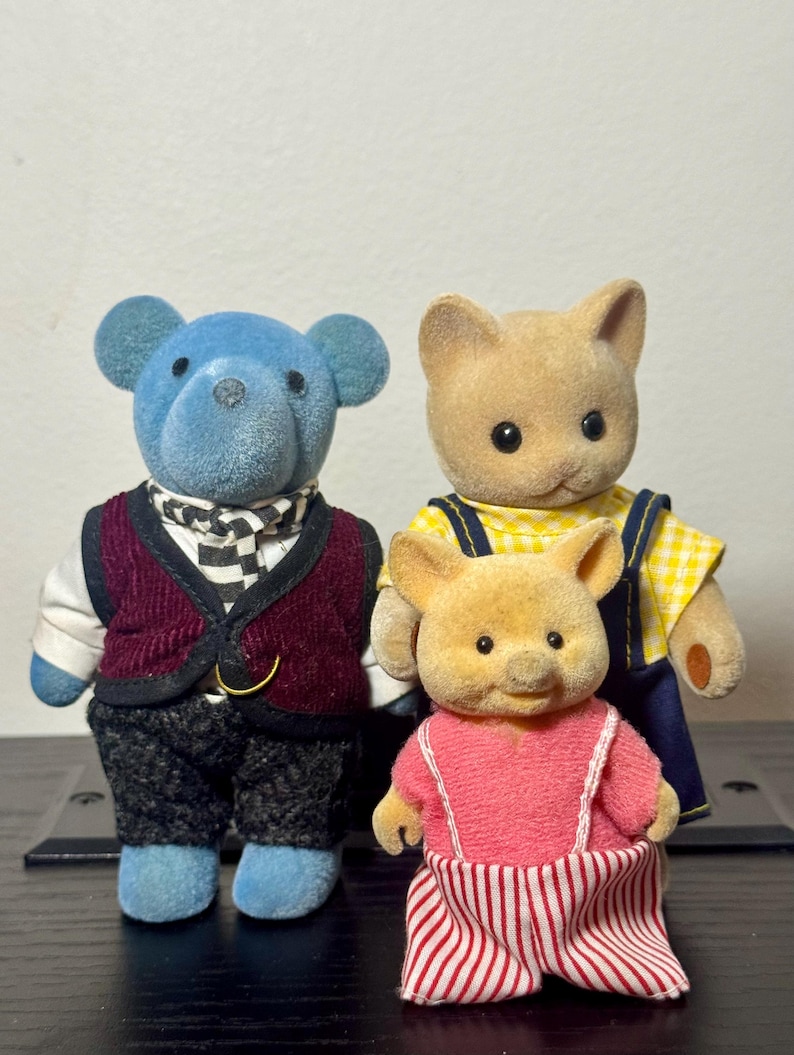 May include: Three miniature animal figurines: a blue bear, a tan cat, and a yellow pig. The bear wears a burgundy vest and checkered scarf. The cat has a yellow top with blue overalls. The pig is in a pink dress with red and white striped pants.