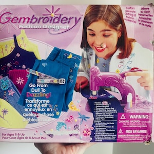 Könnte beinhalten: Eine Gembroidery Fashion Designer-Kit-Box mit einem Mädchen und einer lila Stickmaschine. Die Box zeigt Beispiele für dekorierte Kleidung und Accessoires. Der Text lautet: „Go From Dull To Dazzling!"