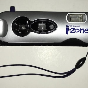 May include: Silver Polaroid i-Zone instant camera with a black base and a dark blue wrist strap. The camera has a flash and a lens. The text "Polaroid i-Zone" is printed in blue on the front of the camera.