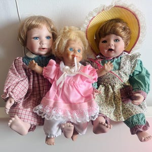 Peut inclure: Trois poupées en porcelaine avec des visages peints et des vêtements détaillés. Les poupées ont des cheveux blonds et bruns, et portent des robes et un chapeau. La poupée au centre porte une robe rose avec une bordure en dentelle blanche.