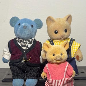 May include: Three flocked animal figurines: a blue bear, a tan cat, and a tan pig. The bear wears a burgundy vest and checkered scarf. The cat has a yellow plaid shirt and blue overalls. The pig wears a pink top and striped pants.