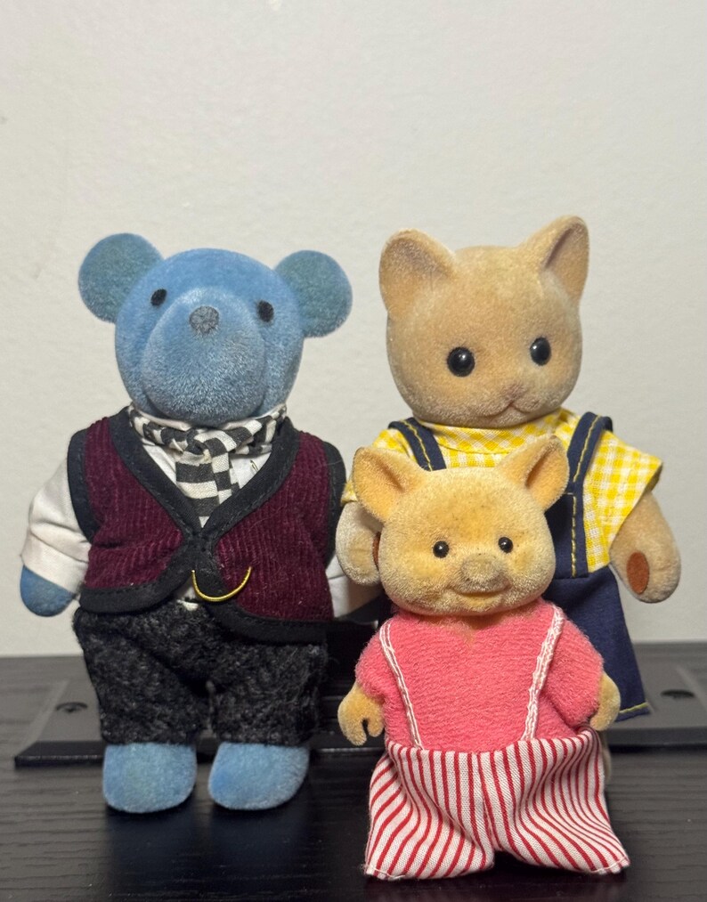 May include: Three flocked animal figurines: a blue bear, a tan cat, and a tan pig. The bear wears a burgundy vest and checkered scarf. The cat has a yellow plaid shirt and blue overalls. The pig wears a pink top and striped pants.