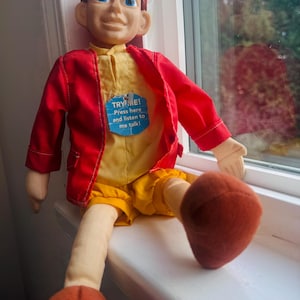May include: A vintage doll with a smiling face, wearing a red jacket, yellow shirt and shorts, and orange shoes. The doll has a tag that reads "TRY ME! Press here and listen to me talk!"